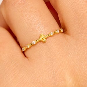 May include: A gold ring with a cross-shaped design, a yellow centre stone, and small white stones. The ring is worn on a finger, highlighting its delicate details and elegant style.