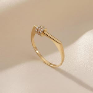 May include: A gold ring with a distinctive design. The ring has a straight, rectangular bar extending from the band, with three small, sparkling bands of gemstones at the top. The ring is set against a soft, cream-coloured background.