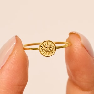 Può includere: Un anello dorato con un ciondolo circolare con un motivo a occhio. La sottile fascia dell'anello sostiene un ciondolo strutturato con raggi di sole attorno all'occhio. L'anello è tenuto tra due dita.
