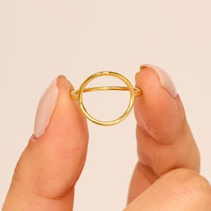May include: A gold-colored ring with a circular design. The ring features a thin band and a prominent open circle in the center. The ring is held between two fingers against a soft, neutral background. The jewelry is simple and elegant.