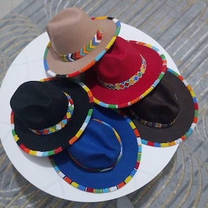 Puede incluir: Una colección de sombreros fedora en varios colores, incluyendo beige, rojo, negro, marrón y azul. Cada sombrero presenta una banda de cuentas coloridas alrededor del borde y un acento decorativo de cuentas en la corona, mostrando un diseño vibrante y artesanal.