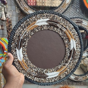May include: Round decorative plate with a brown center and beaded border. The border features a black, braided edge, with white, brown, and gold beads in a feather design. Additional similar plates are visible in the background.