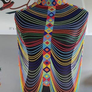 May include: A beaded necklace displayed on a black mannequin. The necklace features a high collar and a central panel with colorful geometric patterns. Strands of beads in various colors cascade down.