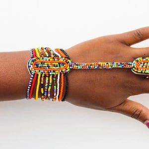 May include: A colorful beaded bracelet and ring set. The bracelet features a large oval design with dangling beads in various colors. The ring has a matching rectangular design, connected by a beaded strand. The jewelry is set against a white background.