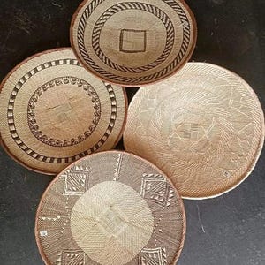 May include: A collection of four round woven baskets in varying sizes and designs. The baskets feature intricate patterns in shades of brown and tan, with decorative borders and central motifs. The natural materials and earthy tones suggest a rustic or bohemian aesthetic.