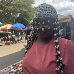 Könnte beinhalten: Eine Frau trägt eine weiße Häkelkopfbedeckung, die mit Muscheln und Perlensträngen verziert ist. Sie trägt ein rosa Kleid. Der Hintergrund zeigt einen Markt mit bunten Sonnenschirmen und verschiedenen Kunsthandwerken.
