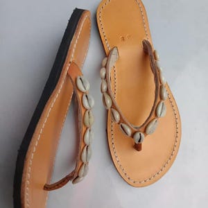 Op de afbeelding: Een paar bruin lederen teenslippers met een zwarte zool. De bandjes zijn versierd met kleine, witte schelpen. De randen zijn gestikt met witte draad en het voetbed is lichtbruin.