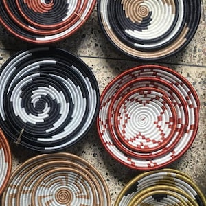 May include: A collection of woven, circular baskets in various sizes and colors. The baskets feature spiral patterns in black, white, brown, red, and gold. The designs are intricate and the baskets appear to be handmade.