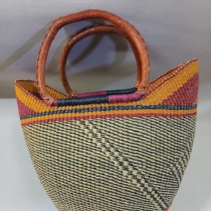 May include: Woven basket bag with a rounded shape and leather handles. The bag features a pattern of black and beige diagonal lines, with bands of yellow, red, and black at the top and bottom. The handles are a light brown colour.