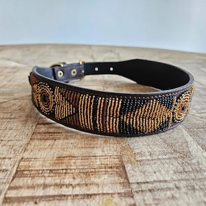Peut inclure: Un collier de chien en cuir marron orné de perles dorées, noires et marron. Le collier est doté d'une boucle en laiton et d'un motif de perles. Le collier est posé sur une surface en bois. La largeur du collier est d'environ 3 cm.