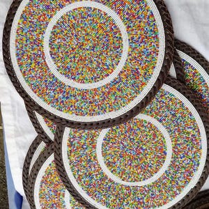 May include: A collection of beaded placemats. Each circular mat features a vibrant, multicolored bead design in concentric circles, with a white border and a dark brown braided edge. The mats are arranged in a layered display.