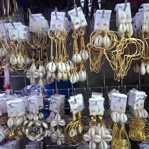 May include: A selection of gold-coloured earrings, many adorned with white seashells. Styles include hoops, dangling designs, and intricate wirework. The earrings are displayed on white cards, suspended from a metal grid.