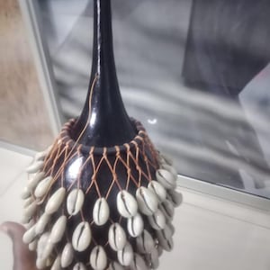 Puede incluir: Un instrumento de percusión negro en forma de calabaza, adornado con conchas blancas y una red tejida. El instrumento tiene un cuello largo y delgado y un grupo de conchas alrededor de la base, con una borla de hilo marrón.
