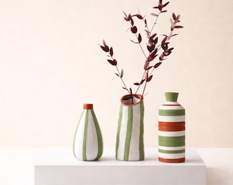 May include: Three ceramic vases with a striped design in green, white, and orange. The vases are arranged on a white surface, with a sprig of burgundy-colored leaves in the center vase. The vases have a modern, minimalist aesthetic.
