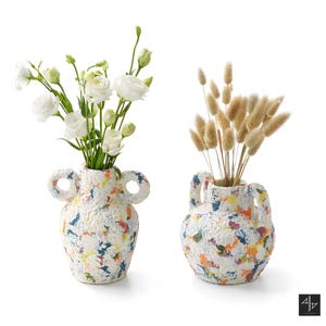 Pode incluir: Dois vasos decorativos com acabamento texturizado e salpicado em branco, azul, amarelo, laranja e vermelho. Um vaso contém flores brancas, o outro capim rabo de coelho seco. Cada vaso tem duas alças.