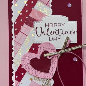 以下が含まれることがあります： ピンクとバーガンディの色使いの手作りバレンタインデーカード。ハート型の装飾、装飾的な紙のストリップ、そして「Happy Valentine's Day」の文字が特徴です。金とピンクのリボンがエレガントさを添えています。