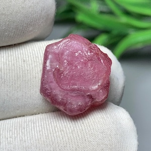 Puede incluir: Una gema rosa en bruto con una estructura cristalina. La gema se sostiene en una tela blanca. El fondo presenta follaje verde. Esta piedra natural es una adición única a cualquier colección.