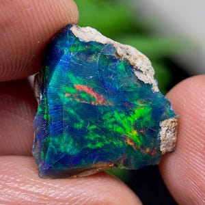 Puede incluir: Una piedra preciosa de ópalo en bruto y sin tallar con un color base azul profundo y negro, con destellos vibrantes de verde neón y naranja. La piedra tiene una forma irregular y dentada con roca de matriz rugosa y clara visible en los bordes.