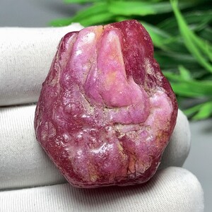 Puede incluir: Una gema de rubí en bruto sin cortar. El rubí es de un color rojo-rosa intenso y saturado, con una superficie texturizada. La piedra tiene una forma natural irregular, con zonas rosadas y beige más claras.