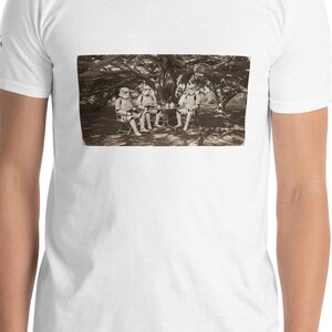 May include: White short-sleeve t-shirt featuring a sepia-toned image of figures in white costumes under a tree. The image is rectangular and centered on the shirt. The shirt is worn by a person.