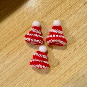 May include: Three miniature red and white striped knit hats with white pom poms. The hats are on a wooden surface.
