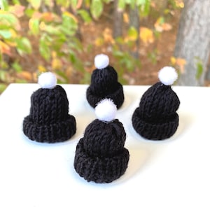 May include: Four black knit hats with white pom-poms. The hats are arranged on a white surface.