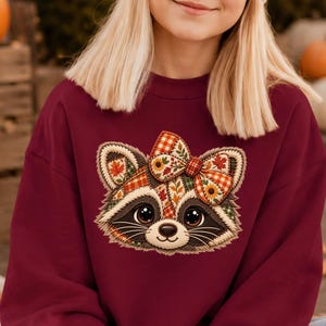 May include: A maroon crewneck sweatshirt featuring a central graphic of a raccoon face with large eyes and a bow on its head. The raccoon is decorated with a colorful autumn-themed pattern of plaid, leaves, and sunflowers in shades of orange and brown.