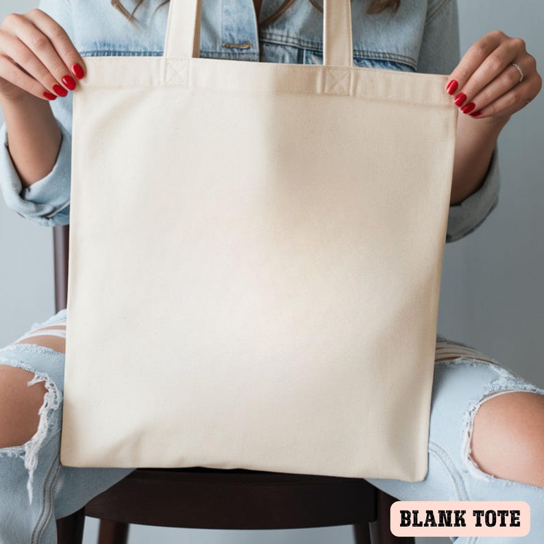 May include: A blank, cream-colored tote bag with sturdy handles. The bag is held up by a person. The text "BLANK TOTE" is displayed in a pink rectangle at the bottom of the image. The bag is made of a durable fabric.
