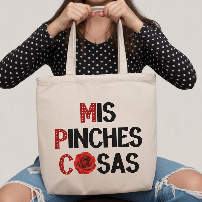 May include: A beige tote bag with the words "MIS PINCHES COSAS" in red and black lettering, and a red rose graphic. The bag has two handles and is held up by a person wearing a black and white polka dot top.