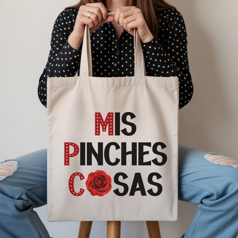 May include: Cream-colored tote bag with the text "MIS PINCHES COSAS" in red and black, featuring a red rose graphic. The bag has two handles and is held by a person wearing a black polka dot shirt and blue jeans.