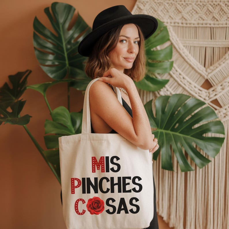 May include: Cream-colored tote bag with the text "MIS PINCHES COSAS" in red and black, featuring a red rose graphic. The bag is held by a person wearing a black hat and top, with large green leaves in the background.