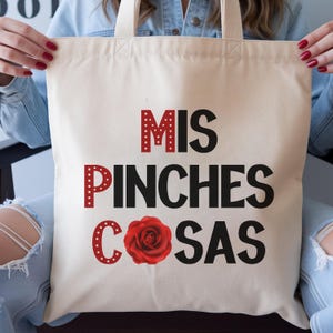 May include: Cream-colored tote bag with the words "MIS PINCHES COSAS" in black and red lettering, and a red rose graphic. The bag has cream-colored handles.