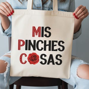 May include: Cream-colored tote bag with the words "MIS PINCHES COSAS" in black and red lettering. A red rose graphic is featured below the text. The bag is held by a person wearing a denim jacket.