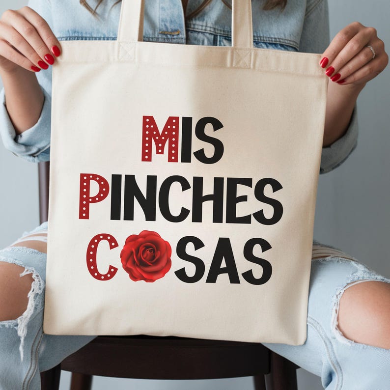 May include: Cream-colored tote bag with the words "MIS PINCHES COSAS" in black and red lettering. A red rose graphic is featured below the text. The bag is held by a person wearing a denim jacket.