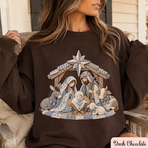 May include: A dark chocolate brown crewneck sweatshirt featuring a printed graphic of a nativity scene. The design depicts a manger with a star above, Mary, Joseph, a baby, a donkey, a cow, and a lamb, all rendered in blue and white gingham patterns.