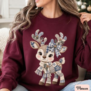 May include: A maroon long-sleeve sweatshirt featuring a central graphic of a reindeer. The reindeer is designed with a patchwork pattern in shades of blue, white, and tan, accented with a blue plaid bow, a matching scarf, and small buttons.