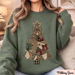 May include: A military green crewneck sweatshirt featuring a large graphic of a Christmas tree made from various patterned fabric patches including leopard print, red and black plaid, and green tartan, accented with several decorative bows.