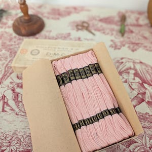Könnte beinhalten: Eine Schachtel mit rosa Stickgarn, wobei der Markenname DMC auf den Spulen zu sehen ist. Das Garn ist ordentlich in einer Pappschachtel angeordnet. Der Hintergrund umfasst einen Stoff mit Vintage-Muster und eine kleine Lampe.