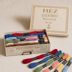 Puede incluir: Una caja de cartón que contiene varias madejas de hilo de bordar MEZ STICKTWIST Mouliné Nr. 25 en varios colores. La tapa muestra la marca, un logotipo de ancla y detalles del producto. Madejas sueltas adicionales están dispuestas delante.