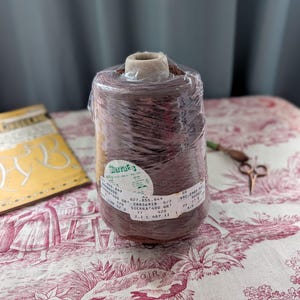 May include: A large spool of brown thread, wrapped in clear plastic, sits on a red and white patterned tablecloth. The thread spool has a label with text and numbers. A pair of small scissors and a yellow booklet are also visible.