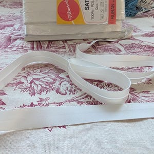 Può includere: Rotoli di sbieco in raso bianco con l'etichetta "Fillawant" e il testo "SATIN 100% POLYESTER Qual. 1939". Diverse lunghezze del nastro sono srotolate su un tessuto fantasia.