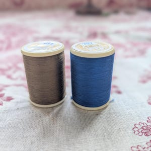 May include: Two spools of thread, one brown and one blue, are displayed. The spools have white bases and gold-coloured tops. The thread is tightly wound around the spools. The background features a floral pattern.