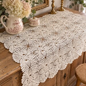 May include: An off-white rectangular crochet table runner featuring a repeating floral motif with intricate openwork stitching and a scalloped edge, draped over a wooden surface with a ceramic pitcher and lidded jar nearby.