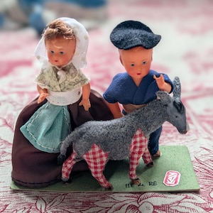May include: A vintage diorama featuring two small dolls dressed in traditional attire, standing with a gray donkey. The donkey has red and white checkered legs. The dolls are on a green base with the text "ILE de RE".