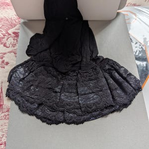 Può includere: Gonna in pizzo nero con un design a strati. La gonna è realizzata in un tessuto scuro e strutturato e presenta un orlo smerlato. La gonna è abbinata a collant neri.
