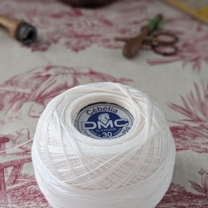 May include: A white ball of DMC Cébélia crochet thread, with a blue and white label. The thread is tightly wound, showing the texture of the cotton. The label indicates it is size 30, made in France, and weighs 75g. The background is a floral patterned fabric.