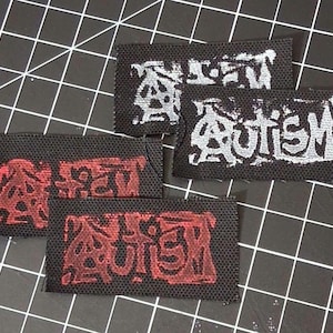 May include: Four black fabric patches with the word "AUTISM" printed in a stylized font. Two patches have red lettering, and two have silver lettering. The patches are rectangular and appear to be iron-on.