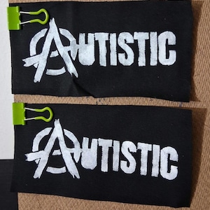 May include: Two black fabric patches with the word "AUTISTIC" in white, stylized with a design resembling a superhero logo. Each patch is held in place by a green binder clip.