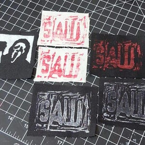 May include: A collection of fabric patches featuring horror-themed designs. One patch displays a black and white image of a character, while others feature the word "SAW" in various colors and styles, including red and white.
