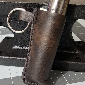 May include: A brown leather lighter case with a metal keyring attached. The case is stitched along the edges and holds a silver lighter. The leather has a distressed appearance.